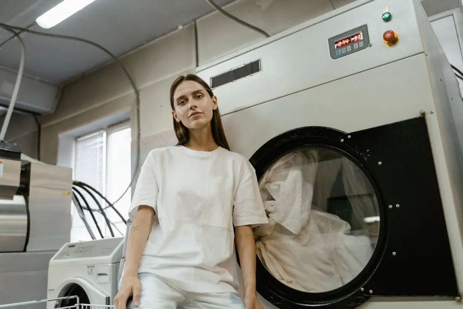 Young woman sitting in a laundry facility beside a large industrial washing machine.